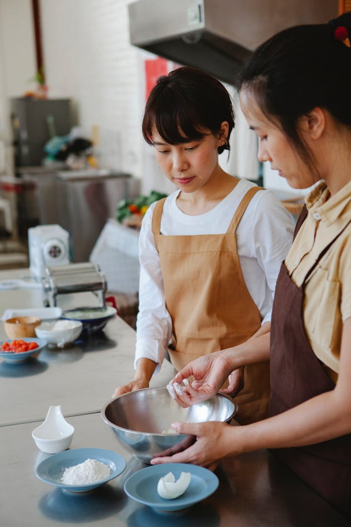 services-02 Two Asian women baking together in a modern kitchen, preparing dough with fresh ingredients.