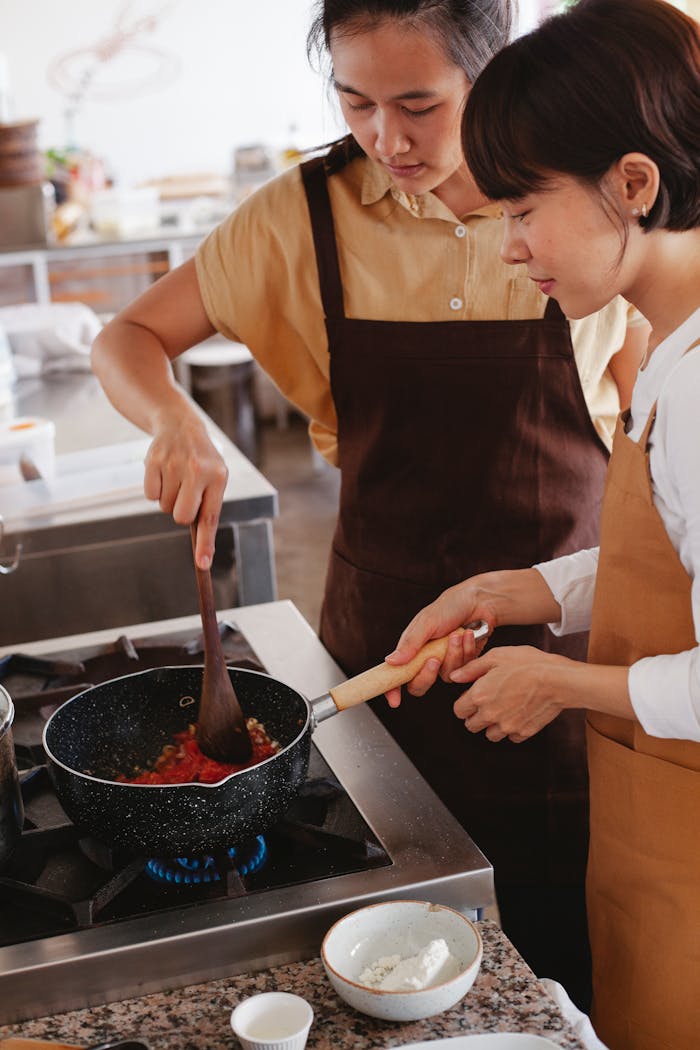 services-03 Two women wearing aprons cook together in a modern kitchen, focusing on a pan dish.
